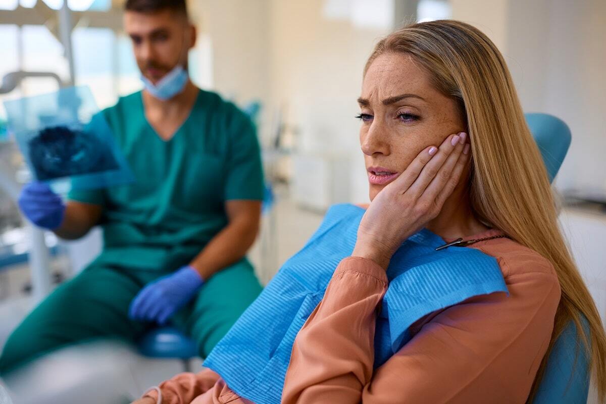 Woman experiencing dental pain holds her cheek while dentist reviews X-rays in treatment room.